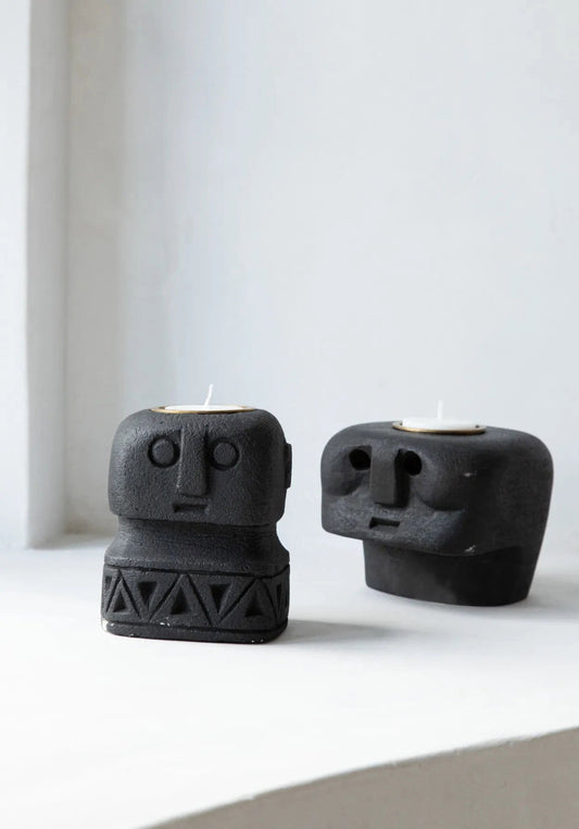De Sumba Stone #26 Kaarsenhouder: two black stone candle holders with carved faces, one with a geometric patterned base.